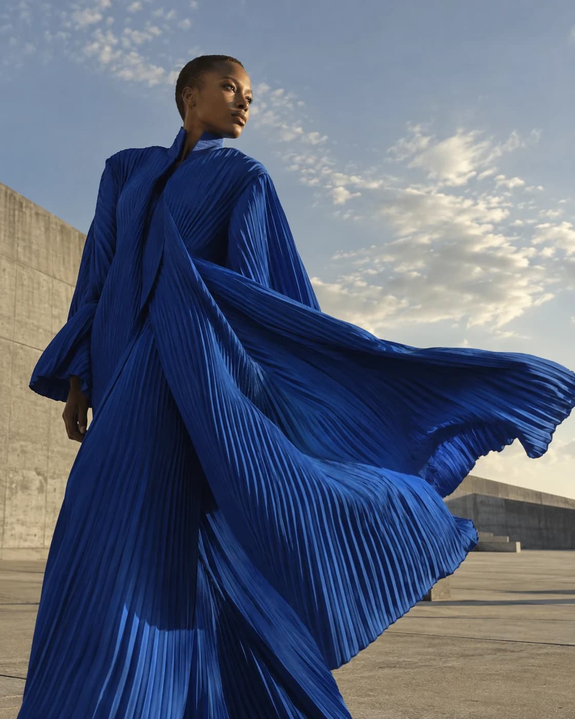 a woman with a close-cropped haircut in a windswept concrete plaza, a low-angle three-quarter frame with the coat sweeping right with hard late-afternoon side light
