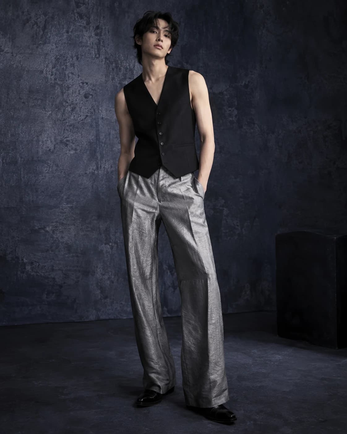 an androgynous model in a black vest and silver-gray trousers in a moody textured studio, a relaxed three-quarter pose with a single softbox with gentle falloff